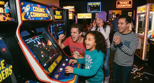 Four people are enjoying a fun time playing an arcade game. Two adults and two children are smiling and laughing as they watch the screen. They are surrounded by other classic arcade games, like Pac-Man and Space Invaders. The Cosmic Commando game they are playing seems to be a hit with everyone.