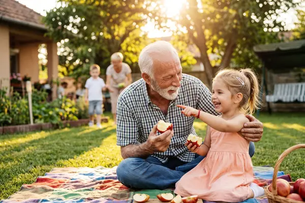 A heart-warming photo showing an older man with white hair, a beard, and a checkered shirt sitting on a picnic blanket on the grass with a young child. They are both holding partially eaten red apples. The man is looking warmly at the girl with a wide smile and has his arm around her. The girl is looking back at him, also with a broad, genuine smile. There is a wooden cutting board with apple slices on the blanket in front of them, along with a wicker basket holding more apples.